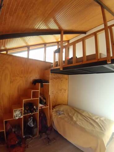 This image shows a cozy wooden room with a loft bed, single bed below, open cubby storage, personal belongings, stairs, and slanted ceiling with warm lighting.