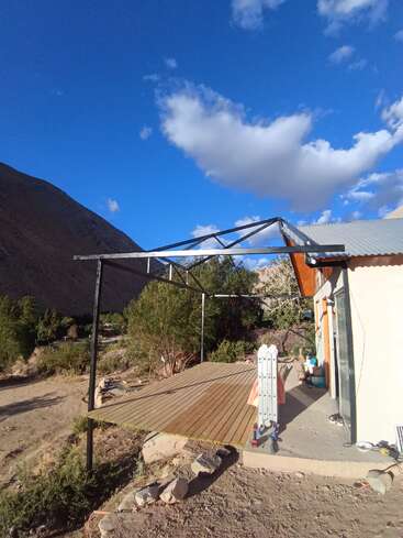 A wooden deck with a metal frame structure sits beside a house in a mountainous, rural area. Blue sky, scattered clouds, and greenery surround the peaceful scene.