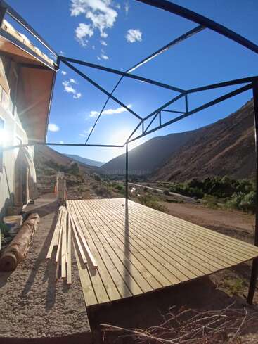 A wooden deck under construction stands beside a house, framed by metal beams. Bright sunlight, blue sky, and mountains create a serene, picturesque outdoor setting.