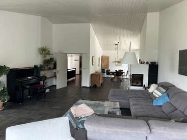 Dieses geräumige Wohnzimmer verfügt über ein graues Sektionssofa, ein schwarzes Klavier, Zimmerpflanzen, einen Kamin, einen Essbereich, hohe Decken, eine moderne Einrichtung und eine gemütliche, einladende Atmosphäre.