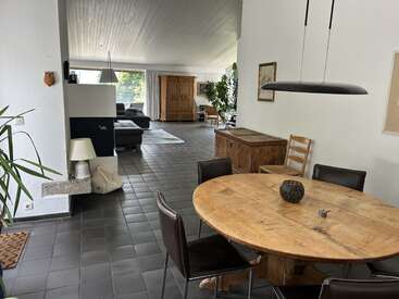 Dies ist ein geräumiger, moderner Wohn- und Essbereich mit einem Holztisch, Lederstühlen, Fliesenboden, großen Fenstern, gemütlichen Sitzgelegenheiten und verschiedenen dekorativen Elementen.