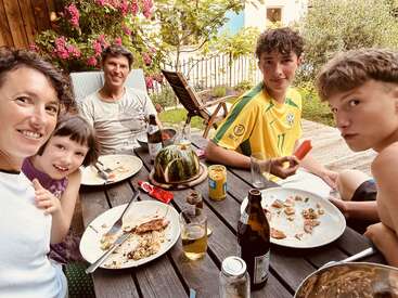 Eine fünfköpfige Familie genießt ein gemeinsames Essen im Freien an einem Holztisch, umgeben von Blumen und Grünzeug, mit Tellern, Getränken und einem Wassermelonen-Mittelstück.