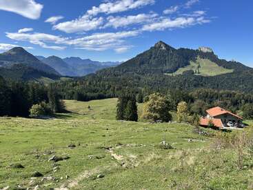 Eine malerische Berglandschaft mit grünen Wiesen, einem bewaldeten Tal, einem Haus mit rotem Dach und dramatischen Gipfeln unter einem strahlend blauen Himmel mit Wolken.