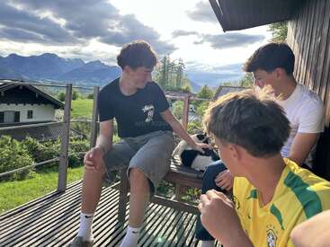 Drei junge Männer entspannen sich auf einer Holzveranda mit malerischen Bergen und Wolken im Hintergrund. Einer streichelt eine schwarz-weiße Katze, während sie gemeinsam die ruhige Atmosphäre im Freien genießen.