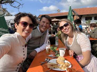 Drei Personen in traditioneller bayerischer Kleidung sitzen im Freien an einem Holztisch, lächeln und genießen Getränke, Kuchen und Sonnenlicht während einer festlichen Zusammenkunft oder Feier.