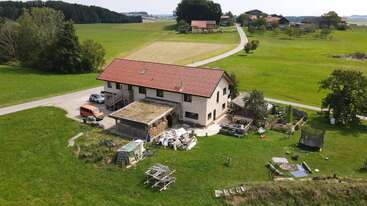 Esta cena rural mostra uma grande casa de fazenda com telhado vermelho, cercada por campos verdes, equipamentos de playground espalhados, uma estrada sinuosa e vários veículos estacionados do lado de fora.