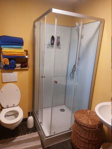 Um banheiro pequeno com chuveiro de vidro, vaso sanitário na parede, cesto de roupa suja de vime, toalhas coloridas dobradas, paredes amarelas, pia branca e tapete listrado no chão.