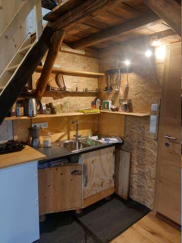 Esta cozinha rústica aconchegante apresenta madeira exposta, prateleiras abertas com pratos, utensílios pendurados, uma chaleira, pia, fogão, armários de madeira e iluminação aconchegante sob uma escada.