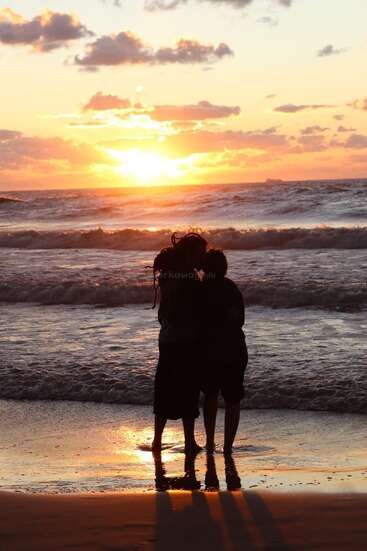 La imagen representa una romántica escena de dos personas abrazadas en la playa al atardecer, con las olas del mar rompiendo suavemente a sus pies.