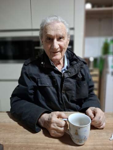 Un anciano de cabello gris está sentado a una mesa de madera, vestido con una chaqueta oscura, sosteniendo una taza y sonriendo cálidamente en una acogedora cocina.