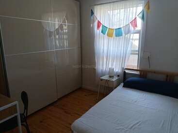 In diesem gemütlichen Schlafzimmer gibt es ein ordentlich gemachtes Bett, einen weißen Kleiderschrank, Holzböden, einen kleinen Nachttisch mit einer Lampe und bunte Wimpel, die über transparenten Vorhängen hängen.