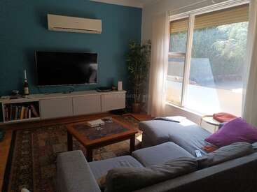 Dieses gemütliche Wohnzimmer verfügt über ein Sektionssofa, ein großes Fenster mit Sonnenlicht, eine blaue Akzentwand, einen montierten Fernseher, eine Zimmerpflanze, ein Bücherregal, einen gemusterten Teppich und einen Couchtisch.