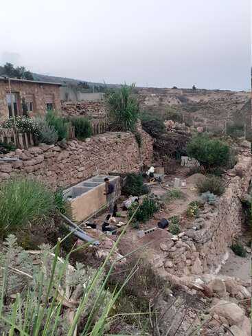 The image depicts a man working on a large sink in a rocky, desert-like area with a stone house and garden, surrounded by a hillside.