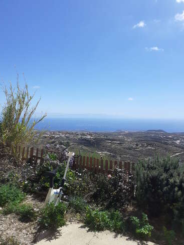 The image depicts a serene garden with a fence, overlooking a vast landscape and the ocean, set against a clear blue sky with a few wispy clouds.