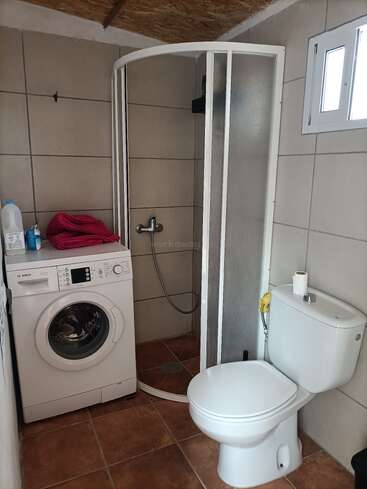 This image shows a small bathroom with a shower, a washing machine, a toilet, tiled walls and floors, some cleaning supplies, and a small window.