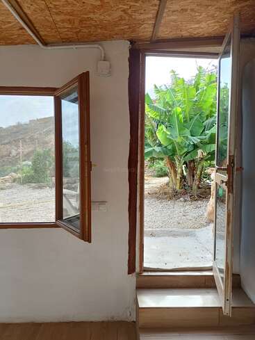 This image shows an open window and door inside a rustic house, revealing green banana plants and a gravel path outside, welcoming fresh air and nature.
