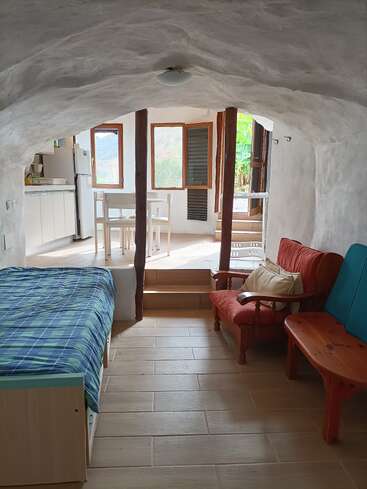 A cozy, rustic cave-like room features a single bed, red cushioned armchair, wooden table, kitchen area, dining table, and bright windows with outdoor views.