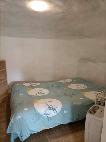 This room has white textured walls, a simple ceiling light, a bed with a blue floral duvet, wooden shelves, tiled floor, and a portable air conditioner.