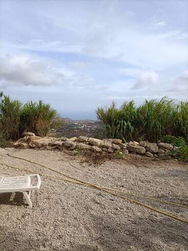 This image shows a gravel ground with a white lounge chair, a stone wall, tall green plants, cloudy sky, and distant hills with a sea view.