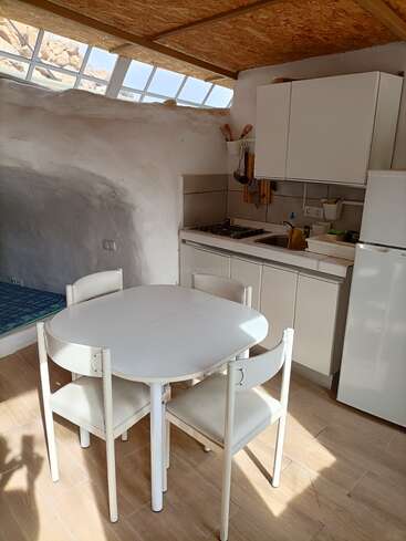A cozy, sunlit kitchen with white cabinets, a small dining table with four chairs, wooden ceiling, tiled floor, and unique curvy white plaster walls and windows.