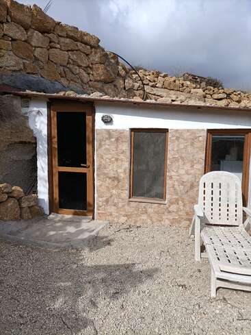 A small stone house built into a rocky hillside features a glass door, two windows, gravel yard, and a white outdoor lounge chair under cloudy skies.