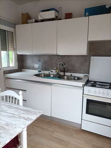 Esta imagem mostra uma cozinha pequena e organizada, com armários brancos, fogão a gás, pia dupla, azulejos cinza na parede atrás da pia, piso de madeira e uma mesa branca de estilo rústico.