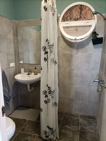 Este banheiro tem paredes e piso revestidos com azulejos cinza, um lavatório pequeno, uma janela redonda, uma cortina de chuveiro com estampa floral, um espelho de parede, toalhas e um chuveiro simples.