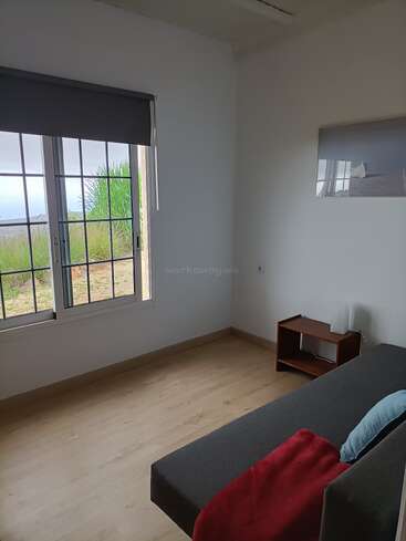 Um quarto minimalista com uma grande janela com vista para a vegetação lá fora, um sofá cinza com uma manta vermelha, uma mesinha de madeira e um quadro na parede.