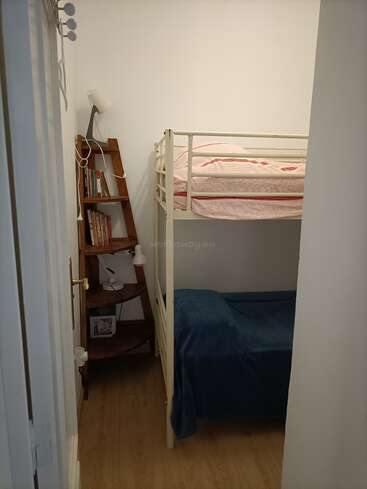 Um quarto pequeno e aconchegante, com paredes brancas, tem uma cama de beliche de metal. Tem uma estante de canto de madeira, livros, um abajur e um cobertor azul na cama de baixo.