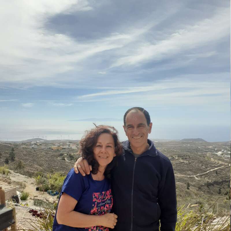 The image depicts a man and woman embracing in front of a scenic landscape, with a cloudy sky and distant hills and buildings visible in the background.