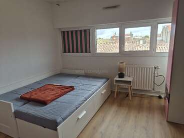 Dies ist ein minimalistisches Schlafzimmer mit einem Einzelbett, blau-grauer Bettwäsche, roter Decke, Holzboden, kleinem Beistelltisch, Lampe und großen Fenstern mit Blick auf die Dächer.