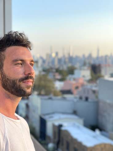 The image depicts a man with dark hair and a beard, wearing a white shirt, standing on a balcony overlooking a cityscape with blurred buildings and a clear sky.