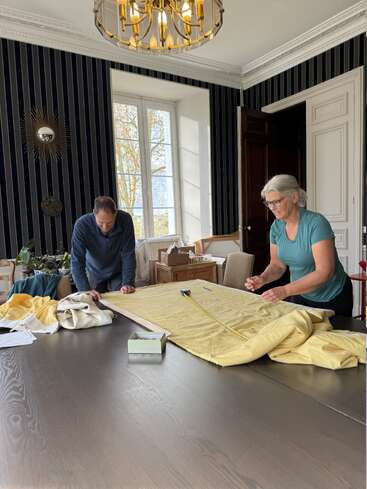Un homme et une femme mesurent ensemble un tissu jaune sur une table dans une pièce élégante avec du papier peint rayé, une grande fenêtre, un lustre et divers objets.