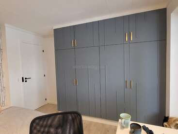 A modern bedroom features a large, blue built-in wardrobe with gold handles, a bed, desk with coffee mug, and a white door with black hardware.