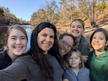 Una sonriente familia de seis miembros se hace un selfie al aire libre cerca de un río. El fondo muestra árboles y cielos despejados, lo que sugiere un agradable día soleado en la naturaleza.