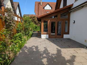 A sunlit patio with stone tiles sits beside a house with large wooden-framed windows and doors. Greenery grows along a wooden fence to the left.