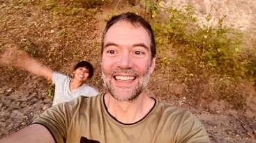 Two people enjoying the outdoors, smiling brightly for a selfie. One person is closer to the camera; the other is joyfully blurred in the background, arms spread.