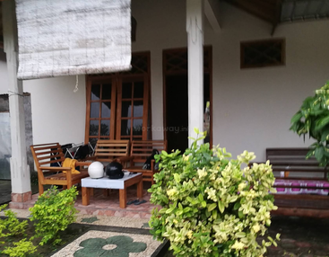 A cozy house porch with wooden chairs, a table holding two helmets, green plants in front, and a shaded window blind hanging slightly above the seating area.
