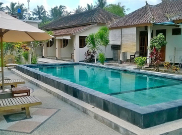 A tranquil outdoor swimming pool surrounded by tropical plants, deck chairs, and umbrellas, with traditional-style cottages and palm trees in the background, under a blue sky.