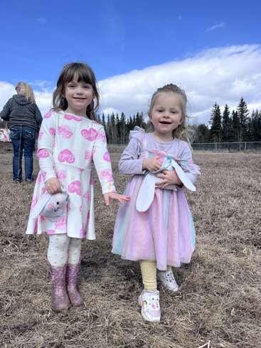 Two young girls smile outdoors on a field, dressed in cute pastel clothing and holding bunny toys. Trees and blue sky with clouds are in the background.