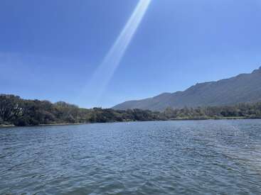 A serene lake reflects the bright blue sky, surrounded by lush green trees and distant mountains, with a single sunbeam shining through the clear atmosphere.