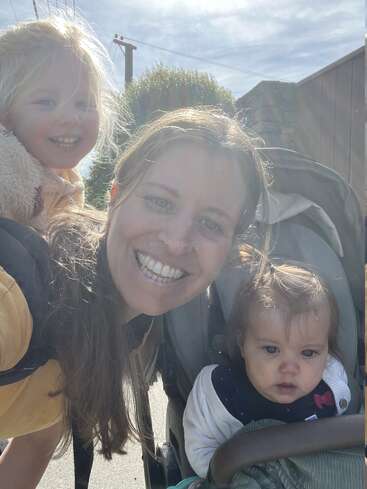 A smiling woman poses outdoors with two young children: one girl on her back, smiling widely, and a baby in a stroller under the bright sun.