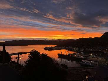 A stunning sunset over a calm harbor, with dramatic orange, yellow, and blue skies reflected in the water. Silhouetted mountains and boats complete the peaceful scene.