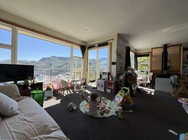 A cozy living room with large windows shows mountain views. A baby plays on a mat surrounded by toys. Sunlight brightens the cheerful, family-friendly space.