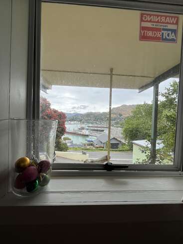 A window view shows a harbor, hills, and houses. A glass vase with colorful ornaments sits on the sill. An ADT Security sign is visible.