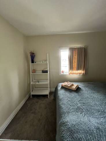 A cozy bedroom with beige walls, a bed covered in a blue blanket, folded towels, a window, curtains, and a white shelf holding various decorative items.