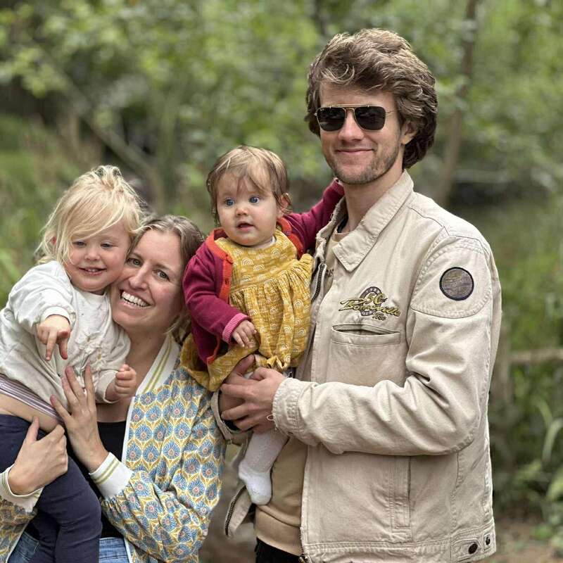 A cheerful family of four stands outdoors. The mother and father are each holding a young child, smiling warmly, surrounded by lush green trees and nature.