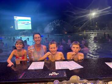 A woman and three children enjoy drinks at a poolside bar at night. People swim in the background, and a large screen displays a race.