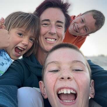 A joyful group selfie featuring a woman and three enthusiastic children, all smiling and laughing outdoors, capturing a moment of fun, happiness, and togetherness.