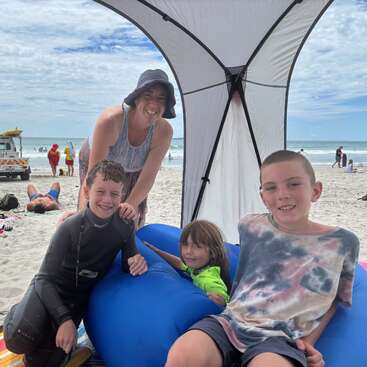A smiling woman with three children relax under a canopy at a sunny beach. The kids are happy, one in a wetsuit, enjoying their summer day.